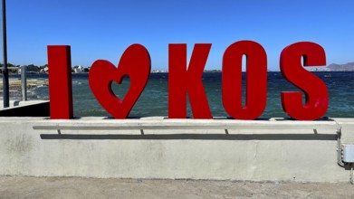 Red lettering 'I??KOS' in front of a blue sea, modern typography style under a clear summer sky,