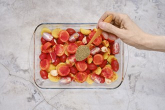 A hand sprinkles oregano over a colorful mixture of tomatoes and other vegetables in a glass baking