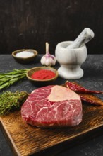 A fresh cut of beef rests on a wooden board, surrounded by garlic, herbs, and various spices. A