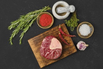 A fresh beef cut rests on a wooden board, surrounded by fresh herbs, spices, and a mortar. The
