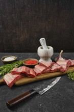 Raw beef ribs are on a wooden cutting board alongside a mortar and pestle. Herbs and spices are