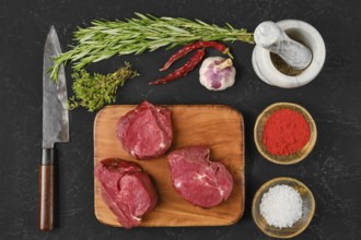 Fresh cuts of beef filet mignon are arranged on a wooden cutting board, surrounded by herbs,