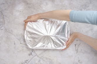 A hand holds a baking dish covered with aluminum foil, ready for the oven. The setting features a
