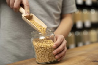 Person filling small glass jar with grains using wooden scoop at bulk store. Concept of zero-waste