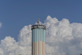 A tall water tower, painted in bright colours, rises against a backdrop of flowing white clouds.