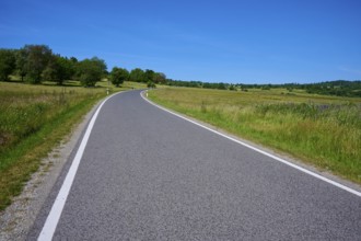 Winding road through a green summer landscape with a blue sky backdrop, Franzosenweg, Lange Rhön,