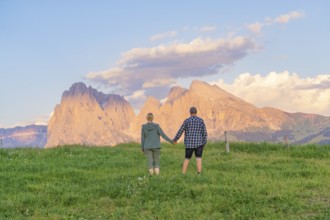 A couple stands hand in hand in a meadow in front of an impressive mountain landscape, Alpe di