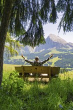 Person with outstretched arms on a bench in front of mountains and fir trees, Alpe di Siusi,