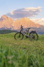 A bicycle stands in a meadow in front of an impressive mountain landscape at sunset, Alpe di Siusi,