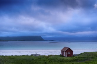 A red wooden hut on the sandy beach of Ramberg (Rambergstranda), sea and mountains. At night at the