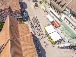 Aerial view of a parade through the centre of a sunny half-timbered town with crowds of people, 950