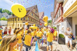 People in yellow T-shirts celebrate in a narrow street with half-timbered houses under colourful