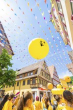 Yellow balloons rise into the sky above a busy street with historical flair and colourful pennants,