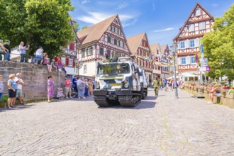 A military vehicle drives on a festive street with half-timbered buildings and people passing by,