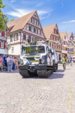 A military vehicle drives past a historic half-timbered backdrop with blue sky and spectators, 950