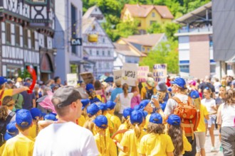 Children in yellow shirts and blue caps march with signs through a busy half-timbered street, 950