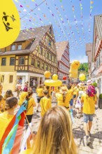 People in yellow clothes walk under colourful pennants in a narrow street in front of traditional