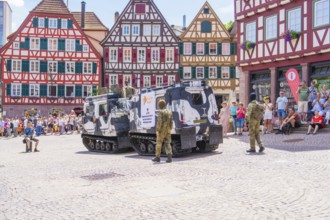 Armoured vehicles stand on a town square with half-timbered buildings and soldiers in camouflage,