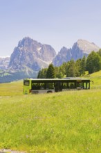 Green bus drives through green meadows in front of majestic mountains in summer, Alpe di Siusi,
