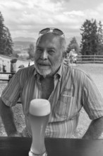 Elderly gentleman with beard and sunglasses in a garden restaurant with a drink, black and white,