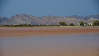 Mountainous landscape with calm lake and small huts under a bright blue sky, salt lake, Tigaki,