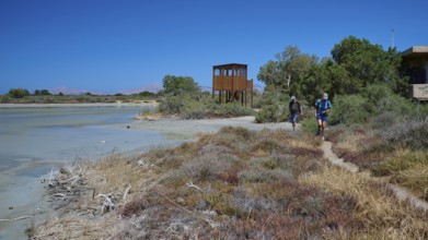 Two hikers on a path next to an observation tower and vegetation, salt lake, Tigaki, nature