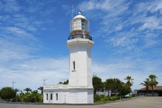 White lighthouse against a blue sky with some clouds surrounded by palm trees and vegetation,
