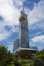 A modern high-rise building rises against a blue sky with clouds. The architecture is futuristic