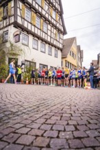 Group of runners on the starting line in front of timber-framed houses, spectators watching the