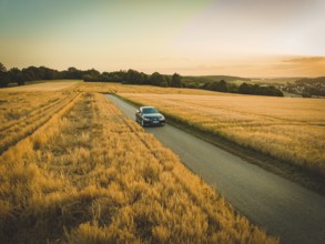 A car drives on a country road between golden fields at sunset, BMW i7, Deer E-Carsharing, Calw,