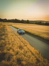 A car drives along a road surrounded by vast fields of corn with a colorful sunset in the