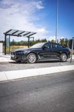 Black car at a modern bus stop under blue sky, BMW i7 electric car, Deer e-Carsharing, Calw,