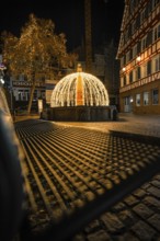 Festively illuminated fountain with lights and shadows on a paved road at night, Calw, Black