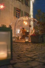 A glass dome with people illuminated at night, next to candles and on a paved path, Gastro Bubbles,