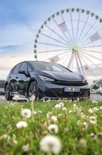 Car in a meadow with Ferris wheel in the background and flowers in the foreground, electric car
