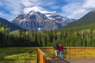 Two tourists standing on a wooden deck are enjoying the breathtaking view of mount robson, the