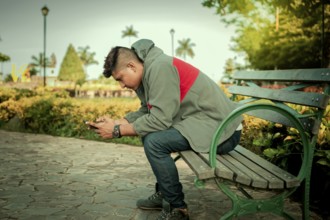 Young man in sweater sitting on a bench texting with phone. Handsome guy sitting on a park bench