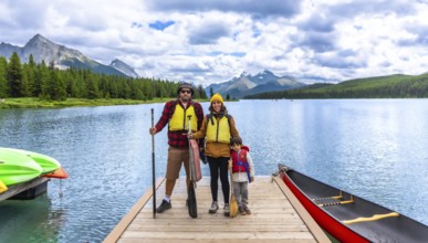 Family wearing life jackets and holding paddles, standing on a wooden dock with canoes and kayaks,