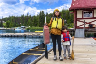 Mother and son holding paddles and wearing life jackets, standing on a wooden dock beside a canoe