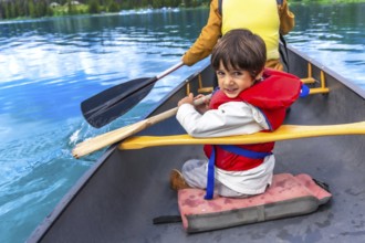 Young boy wearing life jacket paddling canoe on turquoise maligne lake with parent in jasper