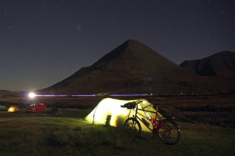 An illuminated tent next to a bicycle in front of a mountain on a clear starry night, Isle of Skye,