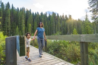 Mother and her son are holding hands and walking along a wooden bridge, surrounded by lush green