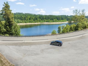A black car drives along a lake, surrounded by a forest, in clear skies, deer e-car sharing, Cupra