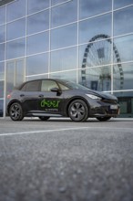 Black car in front of a modern building with glass façade, Ferris wheel reflected in the windows,