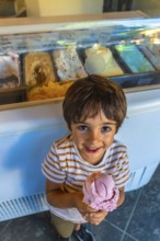 Smiling child holding a strawberry ice cream cone in front of a display case full of colorful ice