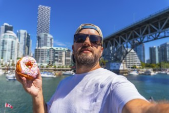 Tourist taking a selfie while holding a donut with burrard bridge and the cityscape of vancouver in