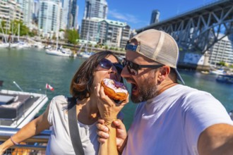 Happy couple biting into a donut together, taking a selfie with vancouver's cityscape and burrard