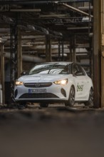 A white Opel with car sharing logo drives through an urban, industrial environment, Deer E-Car