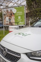 A white Opel with car sharing logo stands in front of an advertising poster and green surroundings,
