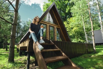 A woman sits on the steps of a wooden cabin surrounded by trees. She holds a drink and looks